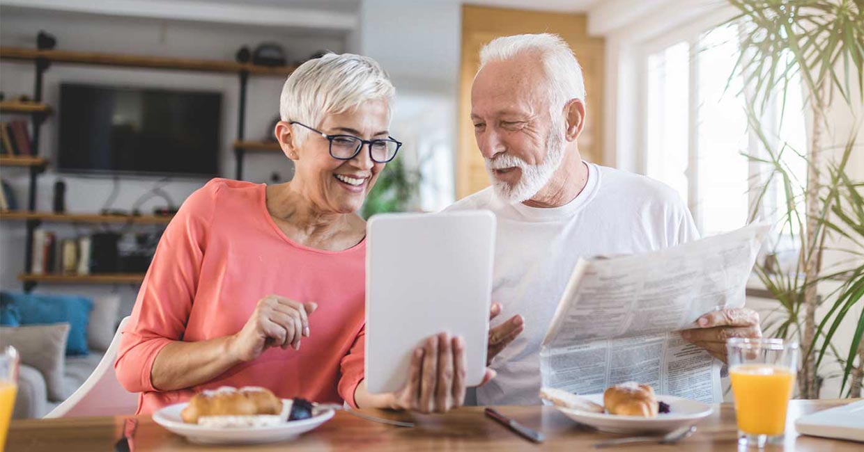 Smiling couple sitting at table, eating breakfast, looking at tablet and having a discussion